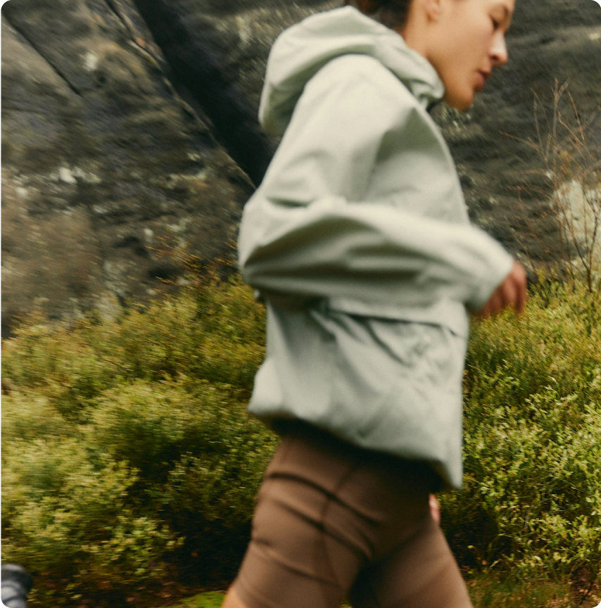 Person in a sage-green jacket jogging outdoors on a trail lined with green shrubs, illustrating active aging and GLP-1 longevity.
